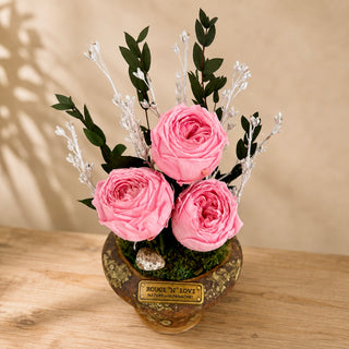 Three pink infinite English roses with preserved greenery and white botanicals in a rustic decorative pot.
