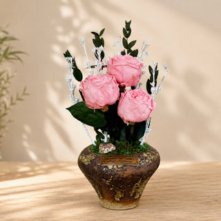 Three pink infinite English roses arranged with preserved botanicals in a rustic decorative pot.